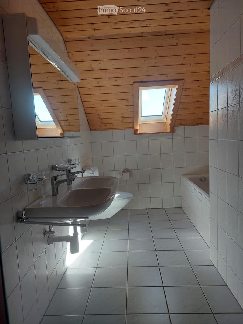Modern bathroom with a double sink, toilet, bathtub, and skylight windows, featuring wooden ceiling and tiled walls and floor.