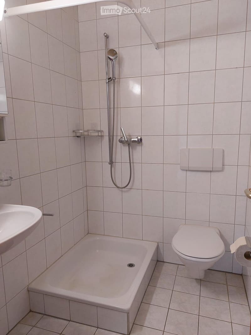 Bathroom with a bathtub, toilet, sink, and a showerhead with a handheld shower attachment. Light-colored tiles cover the walls and floor, and a towel rack is mounted on the wall.