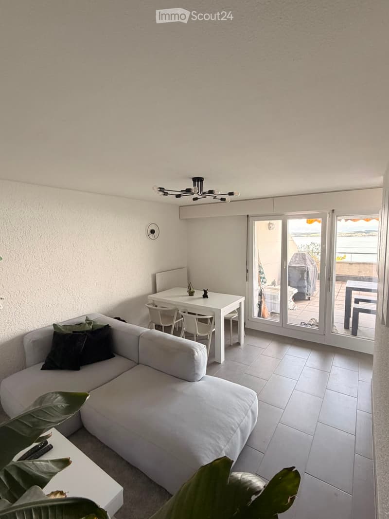 Modern living room with a white sofa, coffee table, dining table, chairs, chandelier, and a view of the sea through large sliding glass doors.