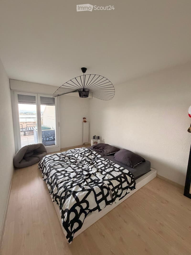 Modern bedroom with a large bed, black and white patterned bedspread, pillows, a modern hanging lamp, and a view through large windows.