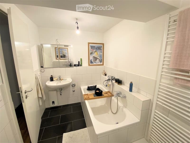 Modern bathroom with white tiles, a sink, bathtub, mirror, and framed picture. Includes a towel and various bathroom accessories.