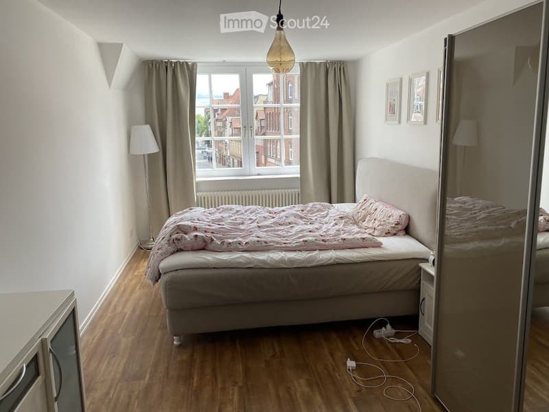 Cozy bedroom with wooden flooring, large window with curtains, bed with patterned bedding, nightstand, mirror, and floor lamp.