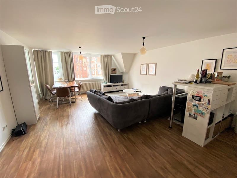 A modern, open-plan living and dining area with wooden flooring, large windows, a grey sectional sofa, a coffee table, a dining table with chairs, and a TV stand.