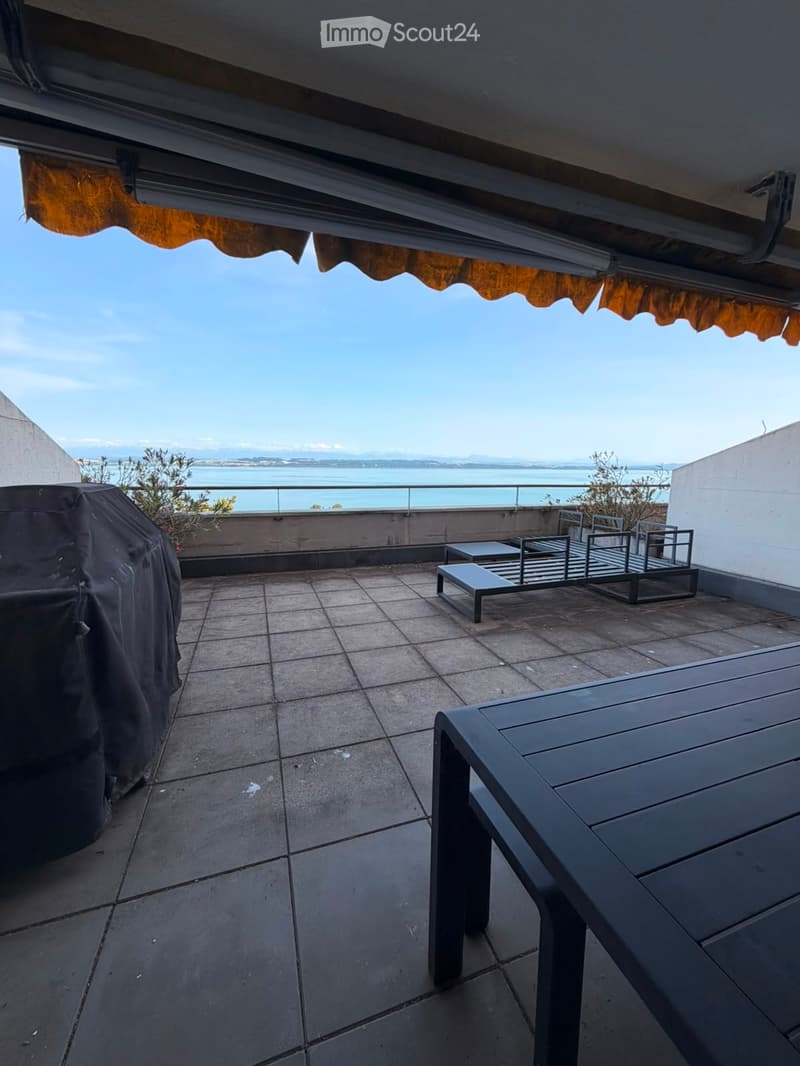 Balcony with tiled floor, outdoor furniture including a table, chairs, and a covered grill, overlooking a scenic body of water.