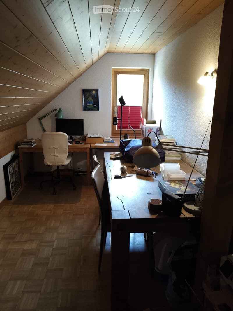 Cozy attic workspace with wooden slanted ceilings, large window with red blinds, two desks with chairs, computer, lamps, books, and framed picture on wall.