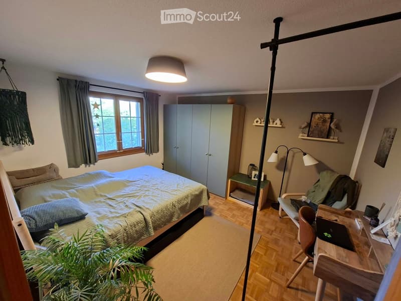 Cozy bedroom with a bed, wooden floor, large window, and a small desk area with a laptop and chair.