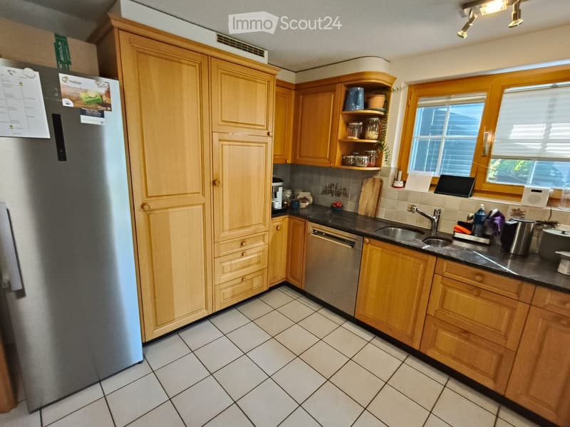 A well-lit kitchen with wooden cabinets, a stainless steel refrigerator, a dishwasher, a sink with a faucet, and various small appliances and items on the countertops.