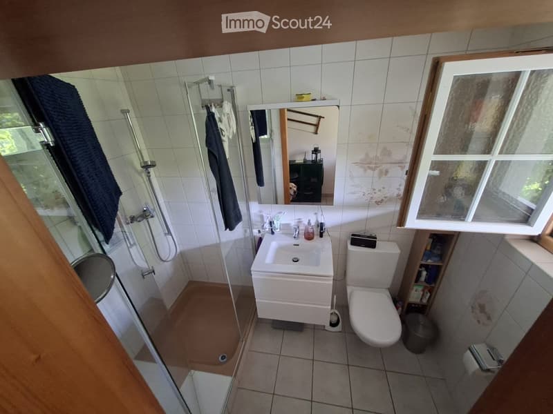 Bathroom with white tiles, a sink, toilet, mirror, shower, and towels. Features a window with frosted glass and a small shelf with toiletries.