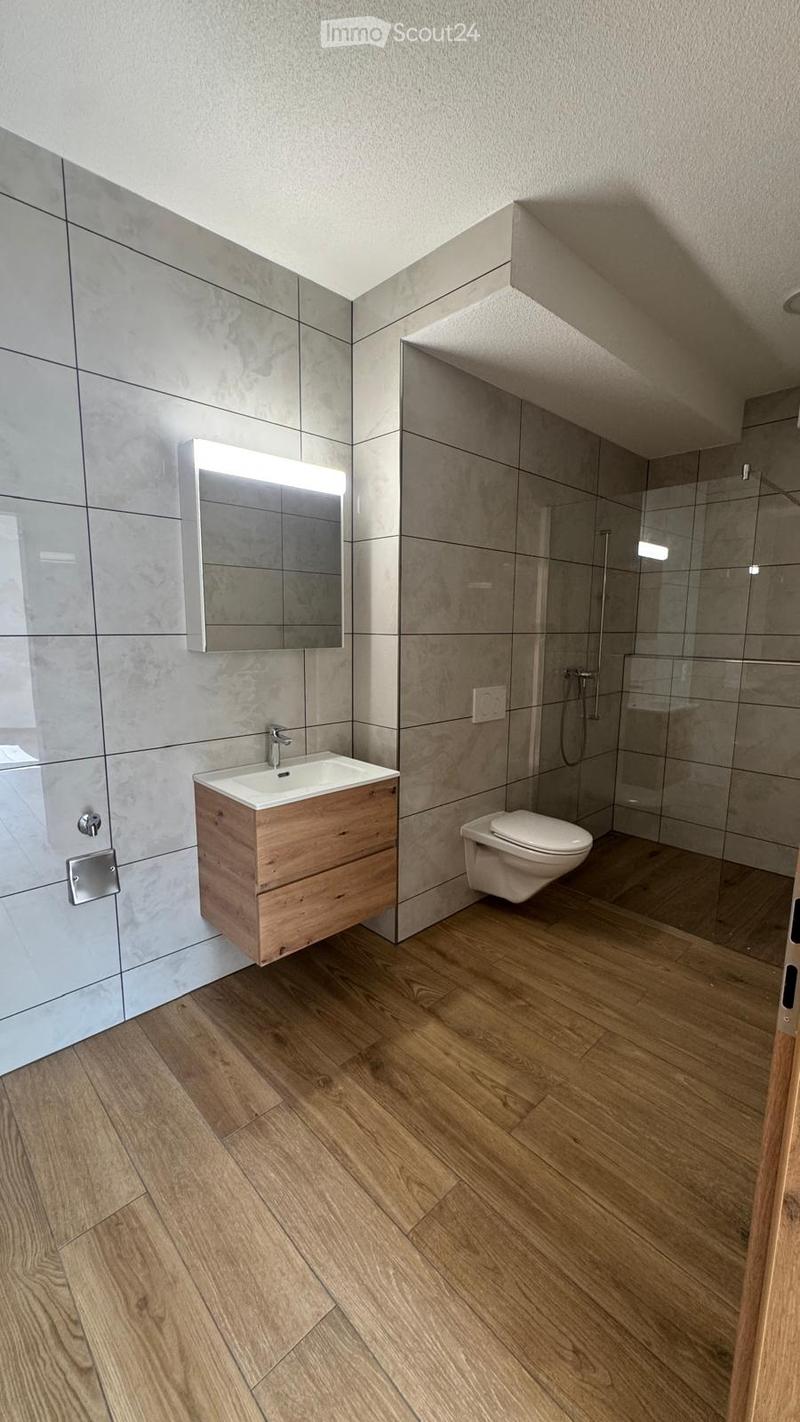 Modern bathroom with wooden flooring, a wooden vanity with a sink, a mirror with built-in lighting, a toilet, and a glass-enclosed shower area.