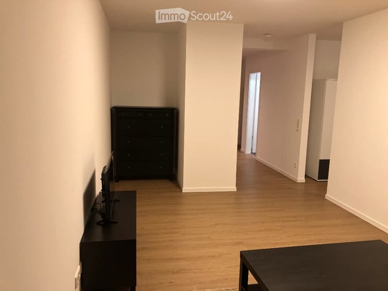 A modern, minimalist living room with light wooden flooring, white walls, and several pieces of black furniture including a TV stand, a small table, and a tall cabinet.