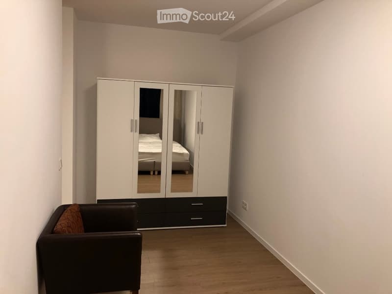 A modern, minimalist bedroom with a large white wardrobe, a black leather chair with a patterned pillow, and a glimpse of a bed in the background.