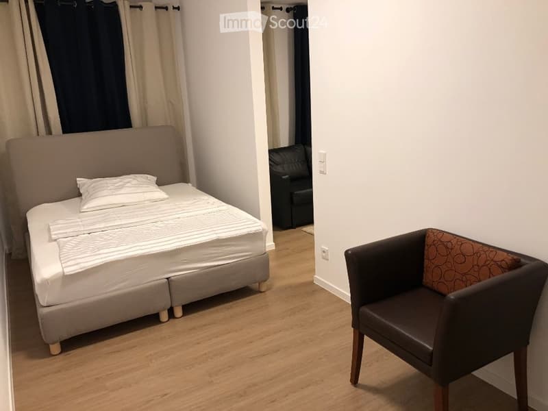 A small, furnished bedroom with a single bed, a leather armchair, and a small sofa in the corner. The room has light wooden flooring and white walls.