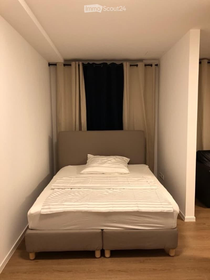 A small, neatly arranged bedroom with a single bed, a headboard, and a pillow. The room features light-colored walls, wooden flooring, and curtains covering the window.