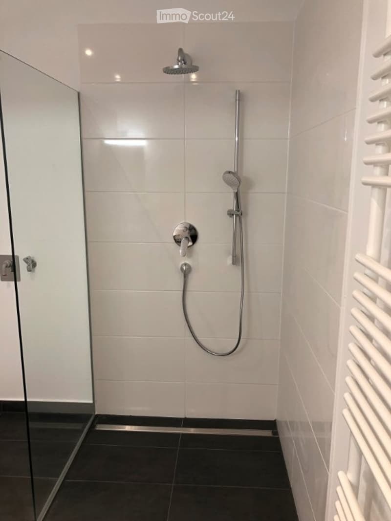 Modern shower area with white tiled walls, black tiled floor, glass door, and multiple showerheads.