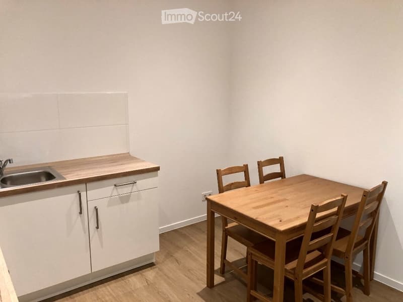 A small, modern kitchen area with white cabinets, a wooden countertop, a sink, and a dining table with four wooden chairs.