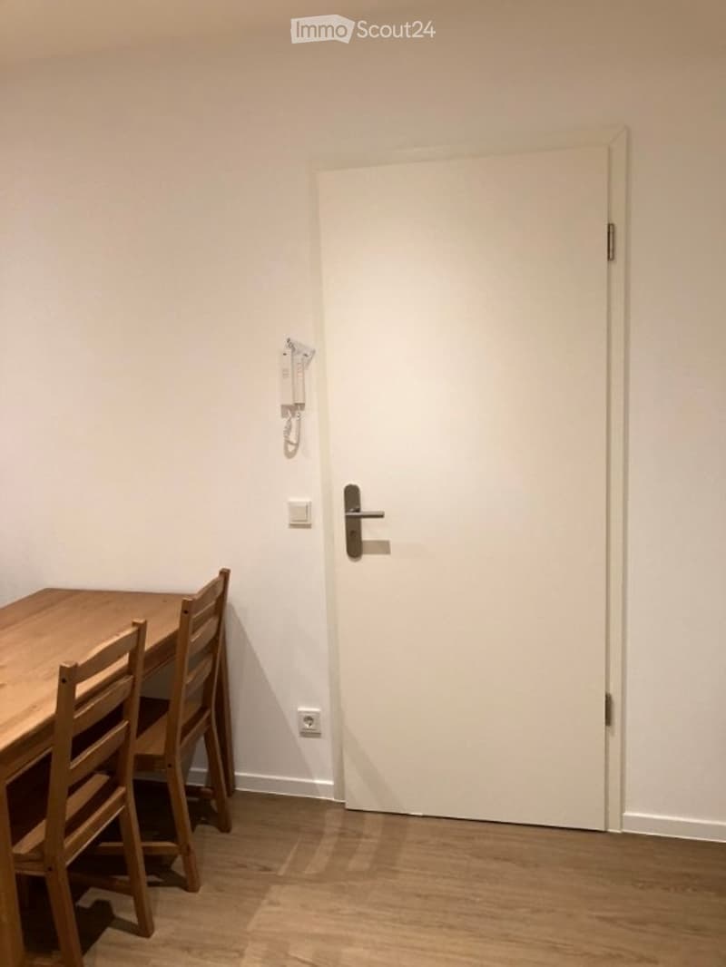 A small, unfurnished dining area with a wooden dining table and chairs, a white door, and a light switch on the wall.