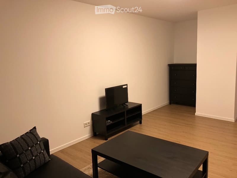 A modern living room with a dark wooden floor, a black sofa with a patterned pillow, a black coffee table, a TV on a stand, and a black drawer unit.