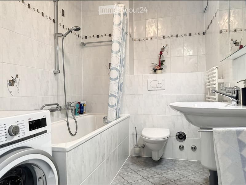 Modern bathroom with white tiles, bathtub, toilet, sink, showerhead, towel rack, and a small flower arrangement on the shelf.