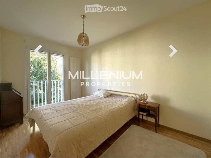 Bright bedroom with wooden flooring, large window leading to a balcony, a neatly made bed, a nightstand with a lamp, and a radiator.