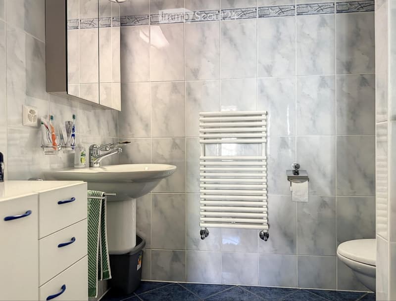 Modern bathroom with a white sink, mirror, radiator, and toilet. The walls are tiled in a marble pattern, and a green towel hangs on a rack.