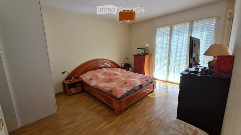 A well-lit bedroom with wooden flooring, a large bed with a floral bedspread, a nightstand with a lamp, a wardrobe, and a table with a screen and decorative items.