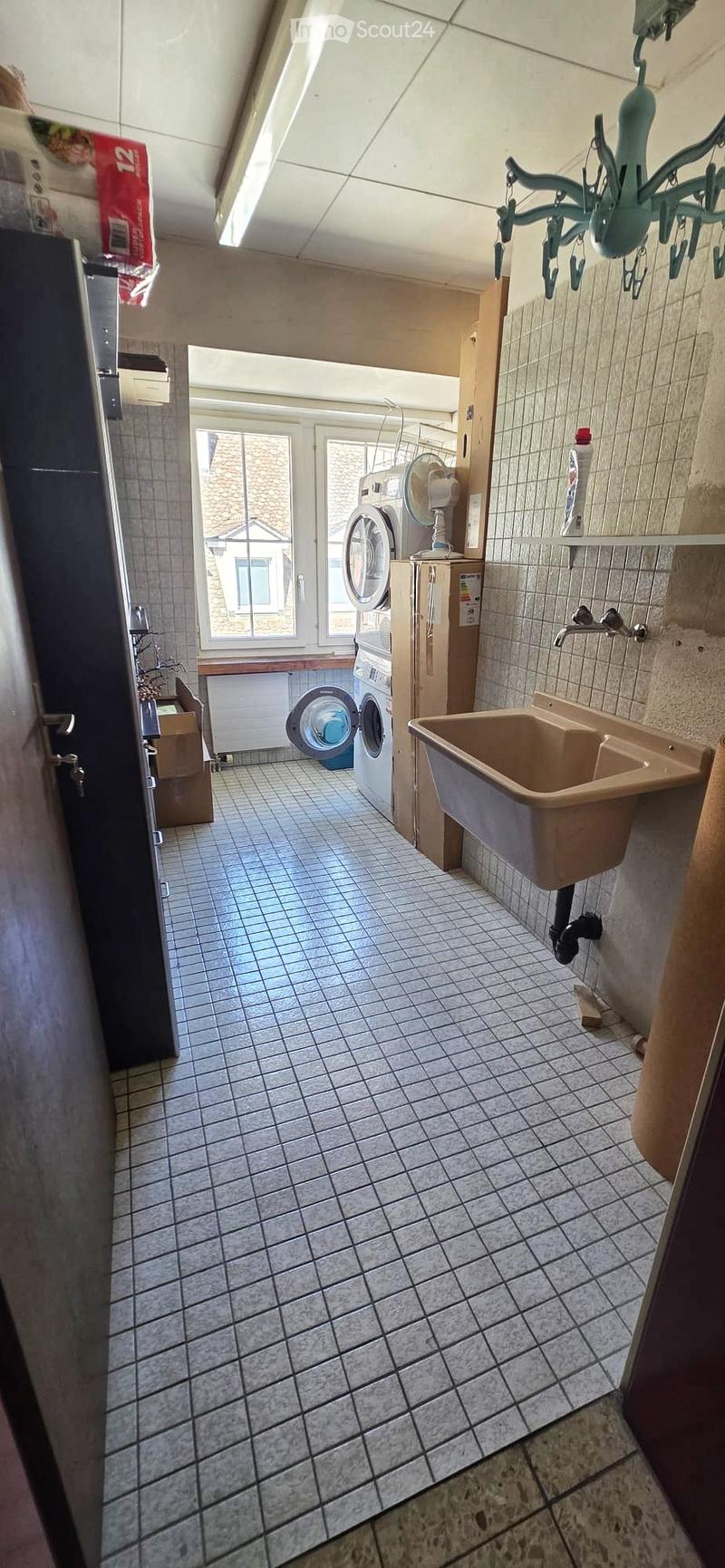 Laundry area with tiled floor, sink, washing machine, fan, and window view.
