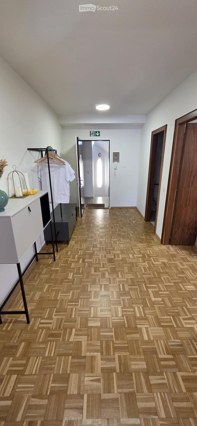 A well-lit hallway with wooden herringbone flooring, white walls, and several doors leading to other rooms. A small console table with a vase and candles is visible on the left side.