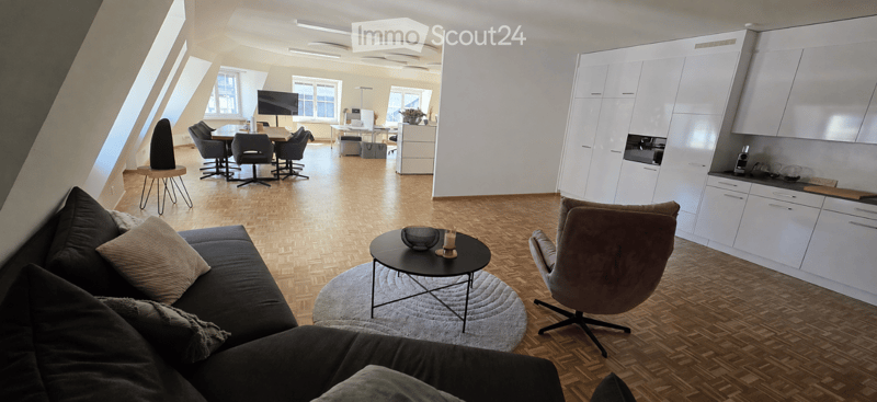 A spacious, modern living room with a dark grey sofa, coffee table, and a round side table. The room features a kitchen area with white cabinets and a dining area with a table and chairs. Large windows allow natural light to fill the space.