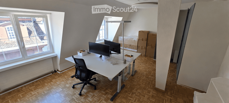 Office space with a modern desk, ergonomic chair, dual monitors, and a standing desk lamp. Wooden flooring and large windows provide natural light.