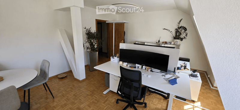 Modern office space with a large desk, ergonomic chair, dual monitors, keyboard, mouse, and a small round table with two chairs. The room features wooden flooring, white walls, and large windows allowing natural light.