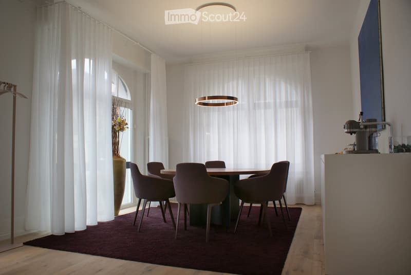 Modern dining area with a round wooden table, four dark-colored chairs, a coffee machine on a counter, and a tall vase with flowers near the window. The space features large white curtains, wooden flooring, and contemporary lighting.