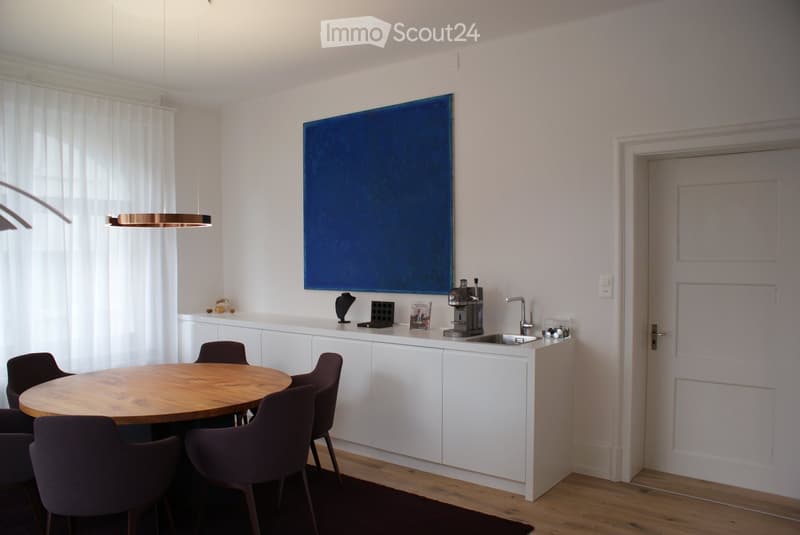 Modern kitchen and dining area with white cabinetry, a round wooden dining table with four chairs, a kitchen island with a sink and coffee machine, and a large blue painting on the wall.