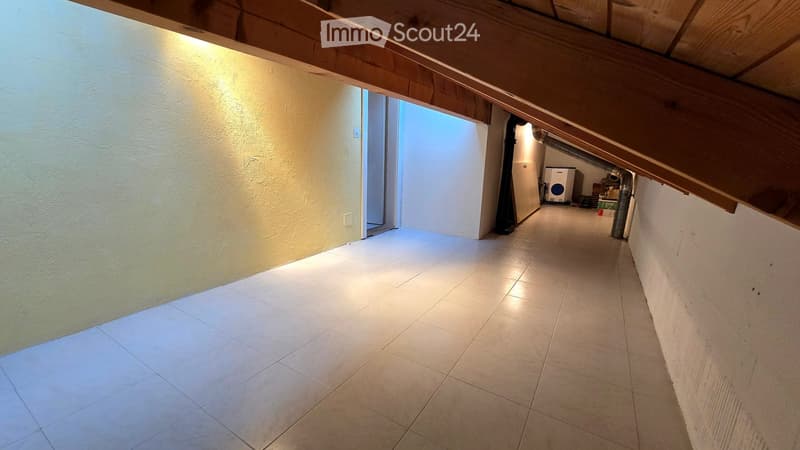 Empty hallway with tiled floor, exposed wooden beams on the ceiling, and a small room visible at the end containing a washing machine and other items.