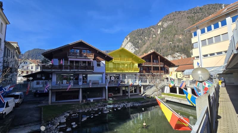 A picturesque town square with colorful buildings, a small canal, and a backdrop of mountains.