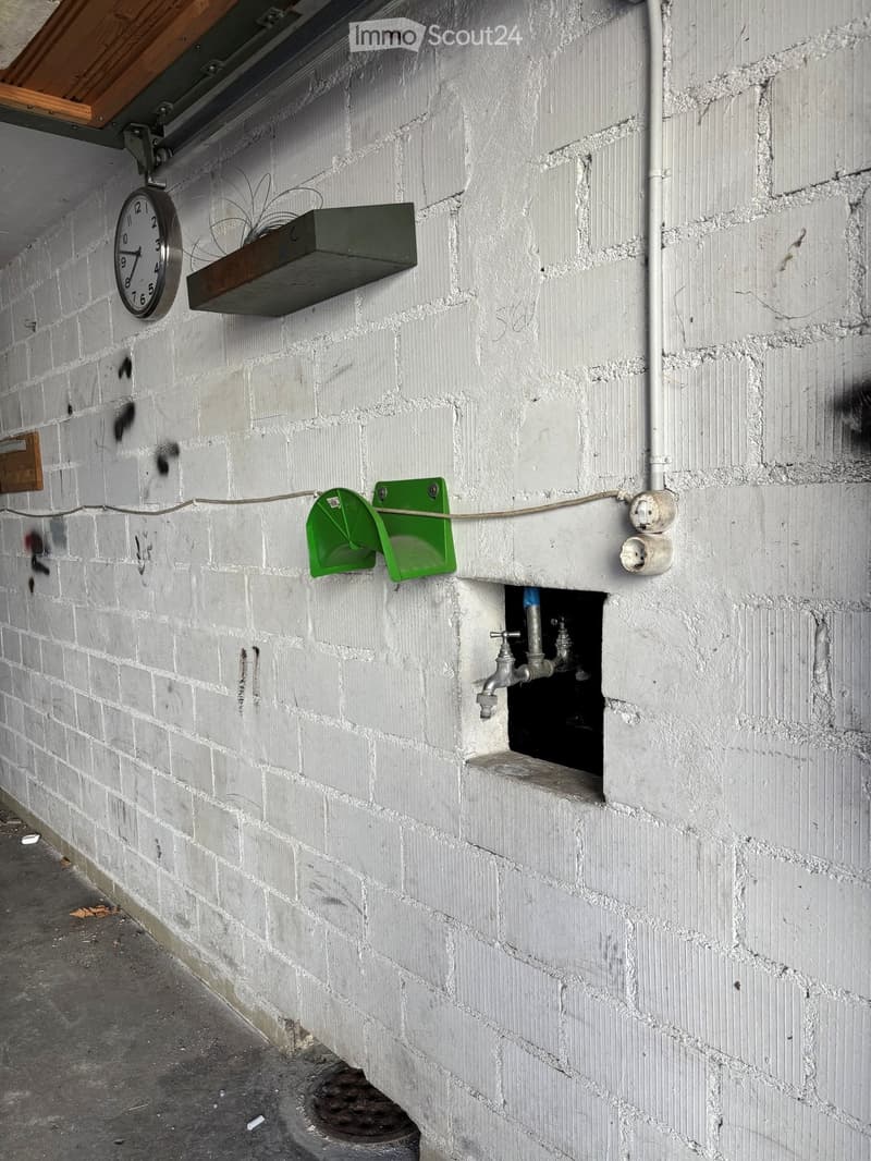 A narrow, gray brick corridor with a clock mounted on the wall, a green metal shelf, and a small faucet protruding from the wall.