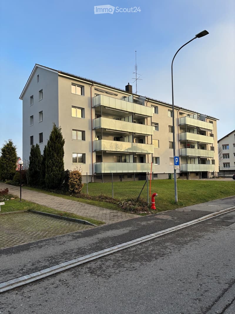 Four-story residential building with multiple balconies, surrounded by greenery and a paved walkway.
