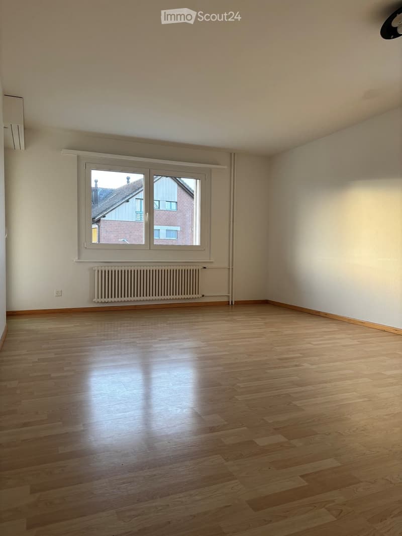 Empty room with wooden flooring, white walls, a radiator, and a large window with white frame showing a view of neighboring buildings.