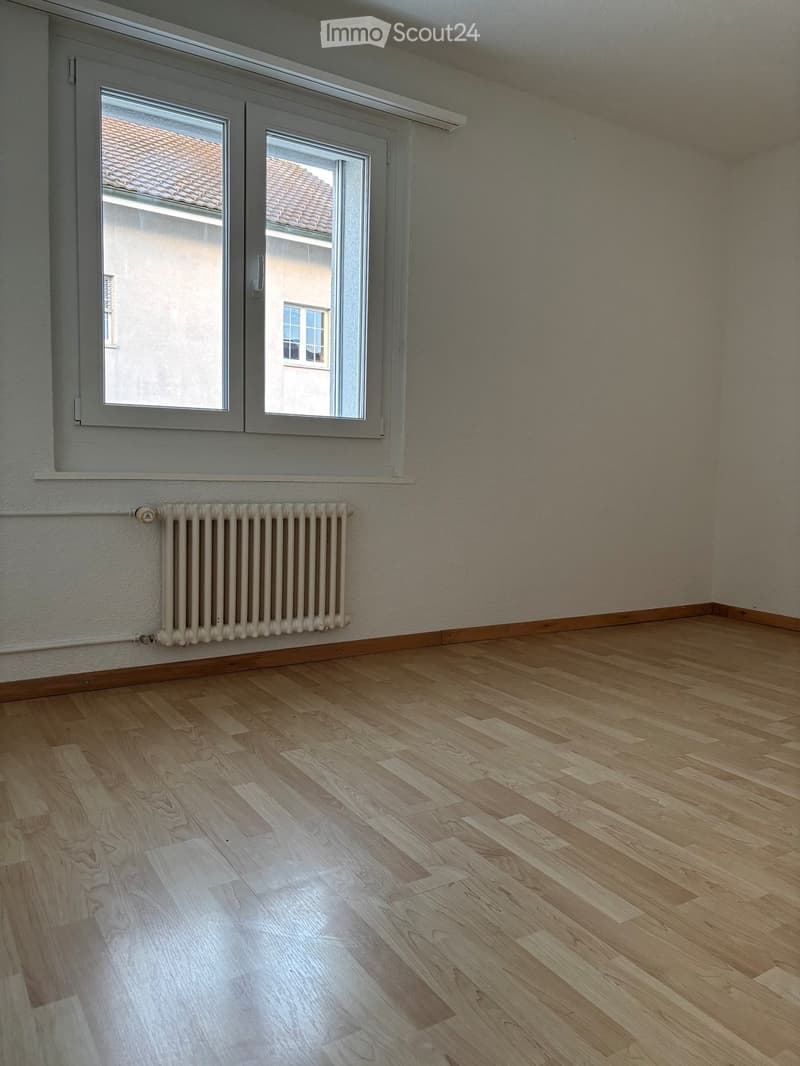 Empty room with wooden flooring, white walls, and large windows with white frames. A radiator is mounted on the wall below the windows.