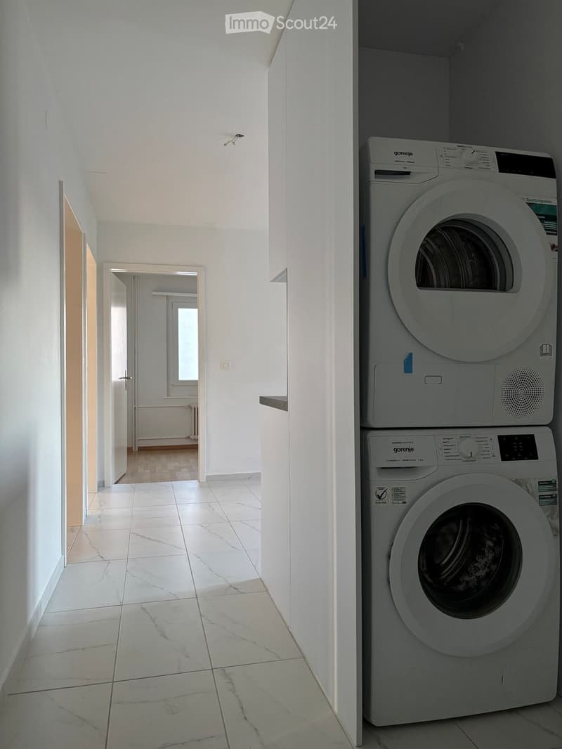 Clean, white-walled hallway with a marble tiled floor, leading to an adjacent room. A stacked washer and dryer unit is positioned on the right side.