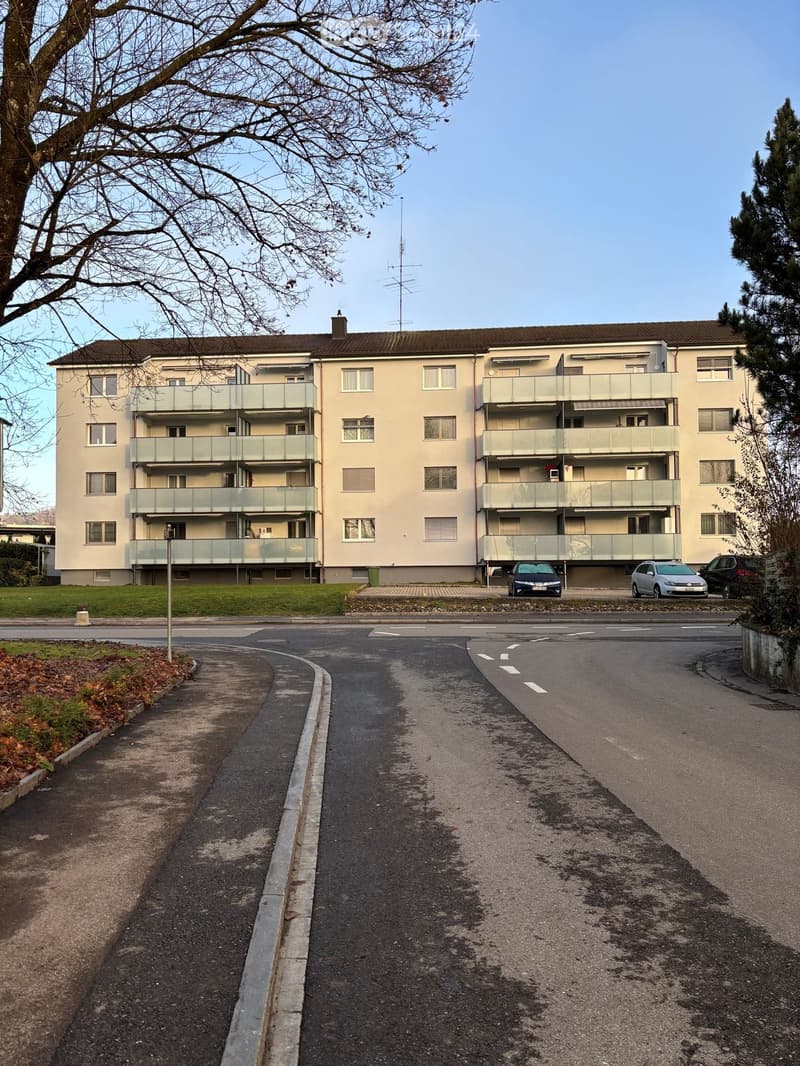 Four-story residential building with multiple balconies and windows, surrounded by a paved road and greenery.