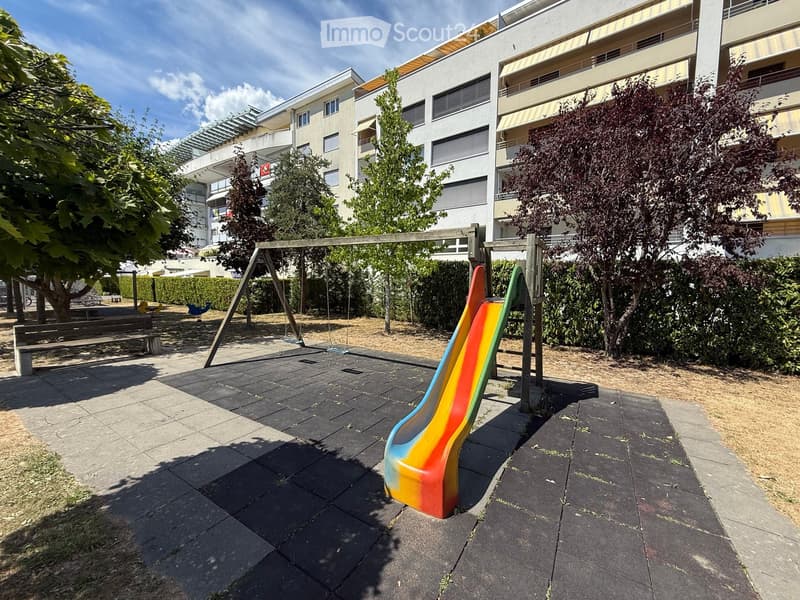 Playground area with a colorful slide, swings, and benches, surrounded by trees and modern apartment buildings.