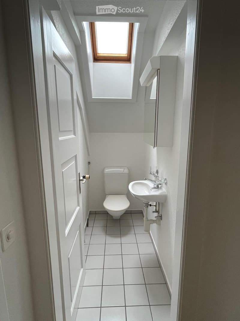 Compact bathroom with white tiled floor and walls, featuring a toilet, sink, and mirror. A small window provides natural light.
