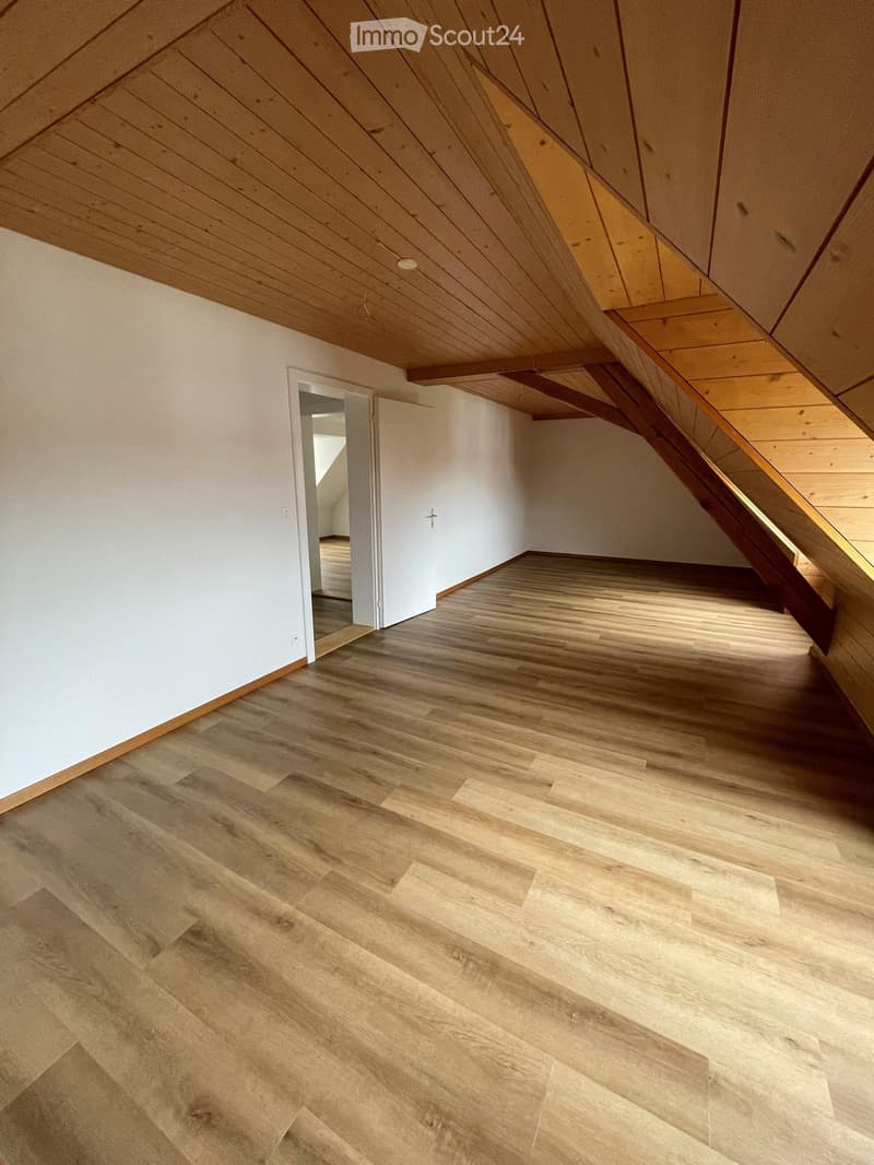 Empty attic space with wooden flooring and slanted wooden ceiling, featuring a doorway leading to another room.