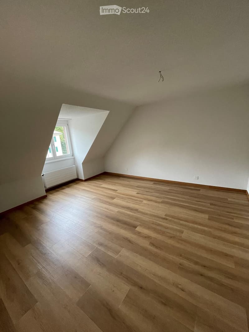 Empty attic room with wooden flooring, slanted ceiling, and a small window letting in natural light. The walls are painted in neutral tones, and there is a radiator visible near the window.