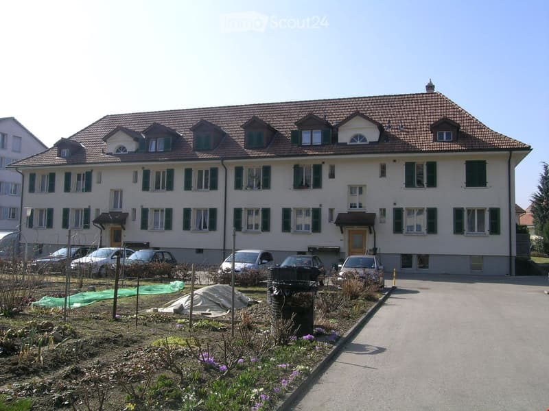 A two-story house with a tiled roof, multiple windows, and a driveway with several parked cars. The front yard has some plants and a small garden area.