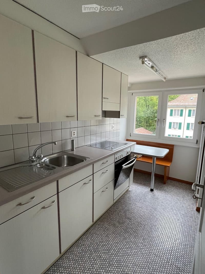 Modern kitchen with white cabinets, stainless steel sink, oven, and a small table with bench seating. The room features hexagonal tiled flooring and large windows offering a view of greenery and neighboring buildings.