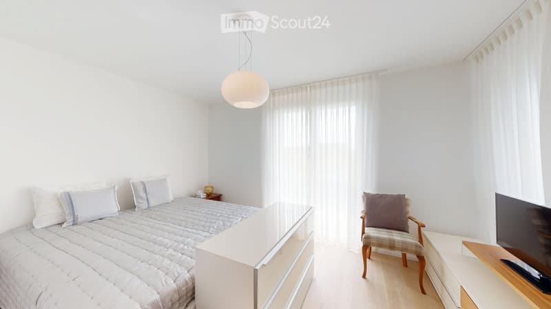Modern bedroom with a double bed, nightstand, armchair, and TV on a wooden stand. The room features white walls, light wooden flooring, and large windows with sheer curtains.