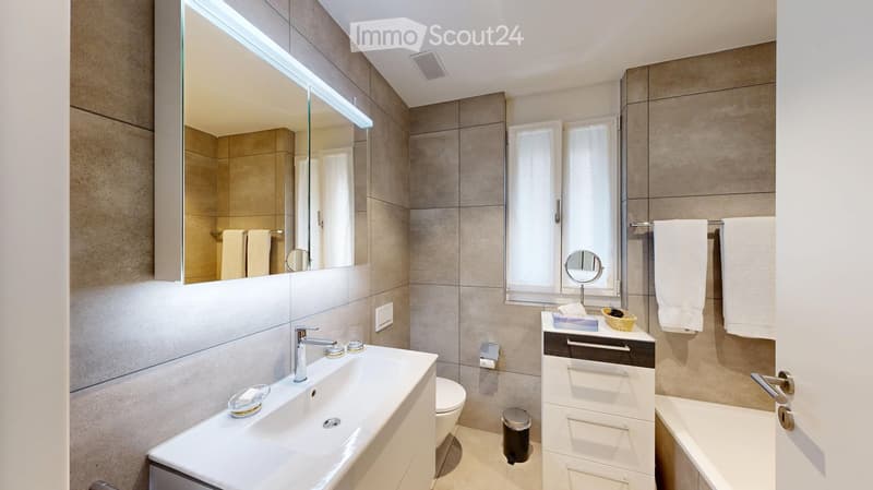Modern bathroom with a large mirror, double sink, bathtub, and towel rack. Features include a sleek faucet, a small basket on the counter, and a handheld mirror on the windowsill.