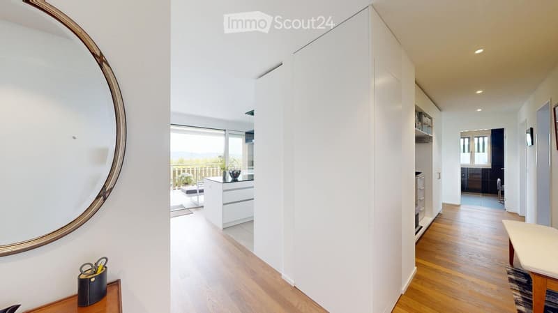 Bright hallway with wooden flooring, large round mirror on the wall, leading to a kitchen area and a balcony with a view.