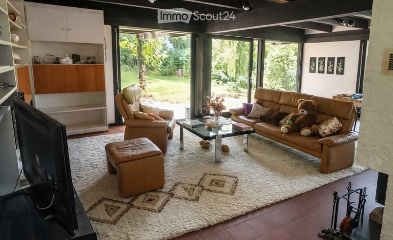 Bright living room with large windows and a view of greenery, featuring a leather sofa, armchair, coffee table, ottoman, and a TV screen.